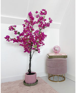 De Kunstplant Bougainvillea 125 cm Paars met levendige paarse bloemen staat naast een roze fluwelen kruk met daarop gestapelde boeken en een roze glazen bol in een minimale, moderne kamer met witte muren.