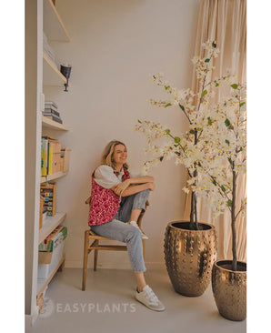 Une personne est assise souriante sur une chaise en bois à côté du bougainvillier artificiel 180 cm blanc dans un pot doré avec texture. À gauche se trouve une étagère avec des livres et de la décoration ; la lumière du jour entre par une fenêtre sur la droite.