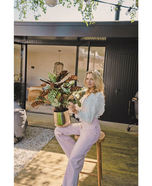 Une femme blonde vêtue d'un chemisier blanc et d'un pantalon violet clair est assise dehors sur un tabouret avec la plante artificielle Calathea Makoyana UV 75 cm en pot rouge, sur une terrasse en bois devant une maison moderne avec des portes vitrées.