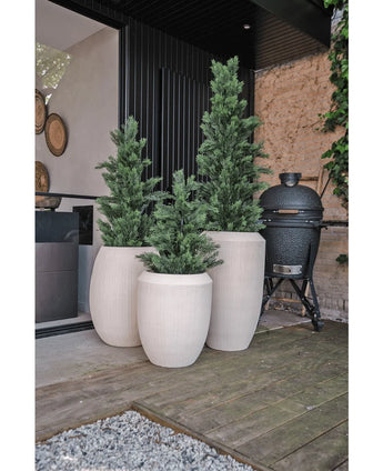 Trois grandes jardinières blanches avec texture et plantes vertes artificielles de conifères artificiels UV 120 cm se dressent sur une terrasse en bois à côté d'une maison moderne. Un grill extérieur noir se trouve à proximité, avec un mur de pierre et de la verdure en arrière-plan.