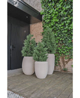 Drie grote beige plantenbakken met Kunstplant Conifeer deluxe UV 150 cm staan op een houten vlonder bij een zwarte deur en klimop muur, waardoor een stijlvolle buiteningang of patio ontstaat voor iedereen die kunstplant tuin inspiratie zoekt.