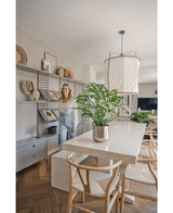 Une femme sourit dans une salle à manger lumineuse et moderne avec des chaises en bois, une table blanche avec la plante artificielle Dieffenbachia de 75 cm comme pièce maîtresse et des étagères ouvertes décorées de livres et d'œuvres d'art.