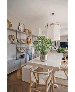 Une femme sourit dans une salle à manger lumineuse et moderne avec des chaises en bois, une table blanche avec la plante artificielle Dieffenbachia de 75 cm comme pièce maîtresse et des étagères ouvertes décorées de livres et d'œuvres d'art.
