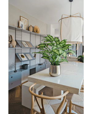 Salle à manger moderne avec une table blanche, des chaises en bois et une plante artificielle Dieffenbachia de 75 cm comme pièce maîtresse. Des étagères avec des livres et des décorations bordent le mur du fond et une grande lampe est suspendue au-dessus de la table.