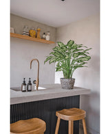 Une cuisine moderne avec un comptoir en béton, deux tabourets en bois et une plante artificielle Dieffenbachia de 75 cm près du robinet en laiton, ainsi que des distributeurs de savon et une étagère en bois avec des pots contre un mur gris clair.