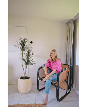Une femme vêtue d'un haut rose vif et d'un jean bleu est assise pieds nus sur une chaise moderne noire et en osier à côté de la plante artificielle Dracaena Fragnans 150 cm dans une pièce lumineuse et minimaliste avec des murs et un sol blancs.