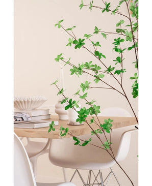 Une salle à manger moderne avec des chaises blanches et une table en bois clair, décorée de livres, de bougies et d'un vase blanc, est encadrée au premier plan par la plante verte à feuilles artificielles Enkianthus de 150 cm.