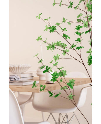 Une salle à manger moderne avec des chaises blanches et une table en bois clair, décorée de livres, de bougies et d'un vase blanc, est encadrée au premier plan par la plante verte à feuilles artificielles Enkianthus de 150 cm.