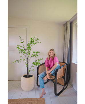 Une femme vêtue d'un haut rose et d'un jean bleu est assise pieds nus sur une chaise en osier moderne à côté de la plante artificielle Enkianthus de 150 cm dans une pièce lumineuse et minimaliste avec des murs blancs et une moquette claire.
