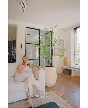 Une femme vêtue de vêtements de détente clairs est assise sur un canapé beige et sourit avec une tasse de café dans un salon moderne avec du parquet, des baies vitrées et l'Enkianthus artificiel de 180 cm comme grande plante en pot.