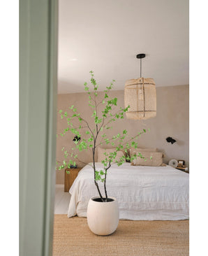 Une chambre minimaliste avec la plante Artificielle Enkianthus 180 cm en grande plante en pot, un lit avec une literie blanche, une suspension tressée, des murs beiges et un tapis en fibres naturelles pour une ambiance simple et aérée.