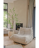 Une chaise longue confortable de couleur crème et texturée se trouve près d’une grande fenêtre et d’une cheminée. A proximité, la plante Artificielle Enkianthus 180 cm ajoute une touche naturelle de vert à la pièce lumineuse, aérée et moderne.