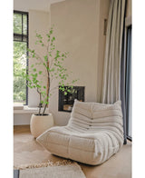 Une confortable chaise longue texturée de couleur crème se trouve à côté de la plante artificielle Enkianthus de 210 cm et d'une cheminée moderne dans un salon lumineux et minimaliste avec du parquet en bois clair et de grandes fenêtres.