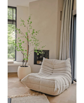 Une confortable chaise longue texturée de couleur crème se trouve à côté de la plante artificielle Enkianthus de 210 cm et d'une cheminée moderne dans un salon lumineux et minimaliste avec du parquet en bois clair et de grandes fenêtres.