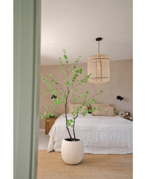 Une chambre minimaliste avec des murs beiges, un lit blanc, un tapis tissé naturel et la plante artificielle Enkianthus 210 cm - une grande plante en pot aux feuilles vertes - au premier plan. Une lampe suspendue tissée est suspendue au plafond.