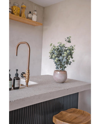 Un plan de travail moderne avec un robinet en bronze, des bouteilles de savon, un tabouret en bois et l'Eucalyptus Artificiel 76 cm. Au fond, des étagères en bois avec des pots et des bouteilles sont montées sur un mur gris clair.