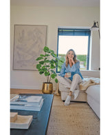 Une femme est assise sur un canapé beige dans un salon moderne et confortable, tenant une tasse et souriante. L'espace est caractérisé par la plante artificielle Ficus de 120 cm dans un pot doré, des œuvres d'art abstraites et la lumière du soleil qui brille à travers les stores bleus.