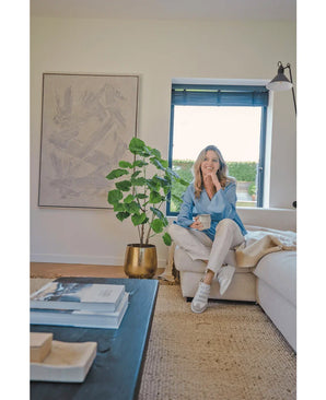 Une femme est assise sur un canapé beige dans un salon moderne et confortable, tenant une tasse et souriante. L'espace est caractérisé par la plante artificielle Ficus de 120 cm dans un pot doré, des œuvres d'art abstraites et la lumière du soleil qui brille à travers les stores bleus.