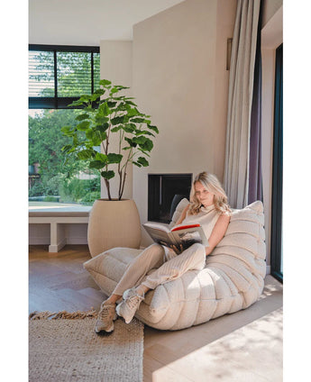 Une femme lit dans un salon lumineux et moderne, assise sur un fauteuil douillet près de la fenêtre, près d'une plante artificielle Ficus 150 cm et de rideaux moelleux, entourée de lumière naturelle.