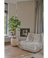 Une confortable chaise longue beige avec structure se trouve à côté de la plante artificielle Ficus de 150 cm dans un salon moderne avec du parquet en bois clair, des murs neutres, une fenêtre et des rideaux gris jusqu'au sol.