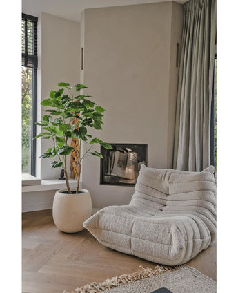 Une confortable chaise longue beige avec structure se trouve à côté de la plante artificielle Ficus de 150 cm dans un salon moderne avec du parquet en bois clair, des murs neutres, une fenêtre et des rideaux gris jusqu'au sol.