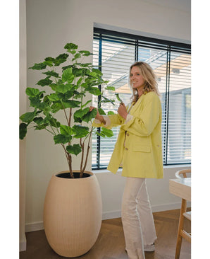 Une femme vêtue d'un blazer jaune et d'un pantalon blanc sourit alors qu'elle s'occupe de la plante artificielle Ficus de 150 cm dans une pièce moderne et bien éclairée avec du parquet et de grandes fenêtres avec stores.
