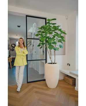 Une femme vêtue d'un blazer jaune et d'un pantalon blanc sourit à côté de la plante artificielle Ficus 180 cm dans une pièce moderne et lumineuse avec parquet et décoration minimaliste.