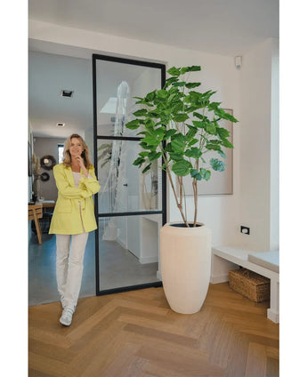 Une femme vêtue d'un blazer jaune et d'un pantalon blanc sourit à côté de la plante artificielle Ficus 180 cm dans une pièce moderne et lumineuse avec parquet et décoration minimaliste.