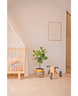 Une chambre de bébé minimaliste avec un cadre de lit en bois clair, un auvent translucide, une plante artificielle Ficus Tobacco Plant Deluxe de 90 cm, des jouets en bois étiquetés Mia et une impression encadrée minimaliste sur un mur blanc uni.
