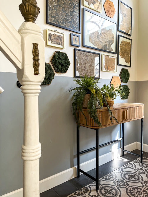 Un hall avec un sol carrelé à motifs, une rampe d'escalier blanche, une console en bois surmontée de la plante artificielle Golden Vine en pot de 25 cm et un mur de galerie décoré d'œuvres d'art encadrées, de cartes et d'accents botaniques.