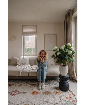 Une femme aux cheveux blonds bouclés est assise sur un canapé beige dans un salon lumineux et pose son menton sur ses mains. À côté d'elle se trouve la plante UV d'hortensia artificielle de 85 cm de haut, éclairée par la lumière à travers une fenêtre aux rideaux beiges.