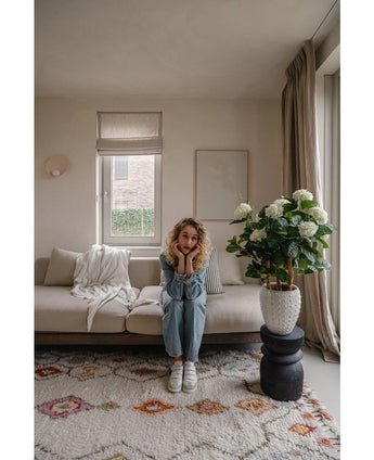 Une femme aux cheveux blonds bouclés est assise sur un canapé beige dans un salon lumineux et pose son menton sur ses mains. À côté d'elle se trouve la plante UV d'hortensia artificielle de 85 cm de haut, éclairée par la lumière à travers une fenêtre aux rideaux beiges.