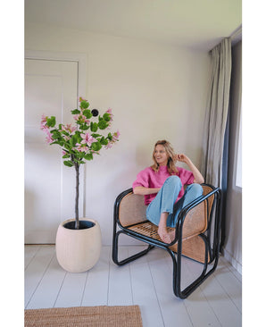 Une femme vêtue d'un pull rose et d'un jean bleu est assise pieds nus sur une chaise en rotin à côté de la plante artificielle Judas 150 cm, un arbre artificiel à fleurs roses, dans une pièce lumineuse et moderne aux murs et au sol blancs.
