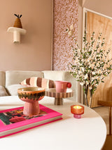 Dans un salon confortable se trouvent une table basse blanche avec un livre rose, un bol, un bol sur pied, une bougie et la plante de cerisier artificielle de 120 cm crème. En arrière-plan se trouve un canapé beige avec des coussins rayés et du papier peint à motifs roses.