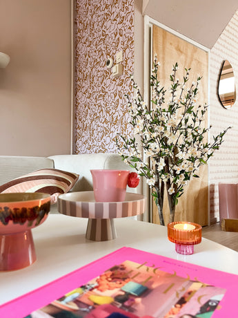 Un salon cosy au décor rose et pastel comprend un plateau rayé sur pied, une tasse rose, une bougie, un bol, un magazine animé et la plante de fleurs de cerisier artificielles 120 cm crème dans un vase haut près d'un mur à motifs et d'une porte en bois.