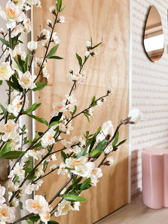 Une plante de cerisier artificielle de 120 cm crème avec des fleurs blanches et rose clair se trouve devant une porte en bois. Derrière se trouvent un tabouret rose, un miroir et du papier peint à motifs roses.