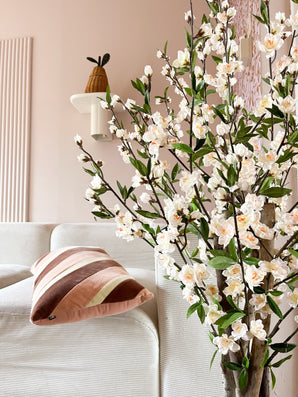 Un salon cosy avec un canapé blanc, un coussin rayé rose et marron et la branche fleurie de cerisier artificiel de 120 cm crème au premier plan. Une plante en pot repose sur une étagère murale contre un mur rose pâle.