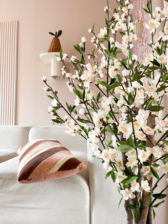 Un salon cosy avec un canapé blanc, un coussin rayé rose et marron et la branche fleurie de cerisier artificiel de 120 cm crème au premier plan. Une plante en pot repose sur une étagère murale contre un mur rose pâle.