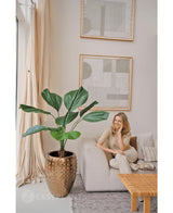 Een vrouw ontspant op een lichte bank in een moderne, neutraal gekleurde woonkamer. Naast haar staat de Kunstplant Lucuala Grandis 120 cm in een pot, terwijl er abstracte kunst boven hangt en natuurlijk licht de ruimte verlicht.