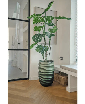 La plante artificielle Monstera 150 cm, avec ses grandes feuilles vertes, se dresse dans un pot au motif ondulé sur un parquet en bois dans une pièce moderne et lumineuse avec une porte vitrée, des œuvres d'art encadrées et un panier en osier à proximité.