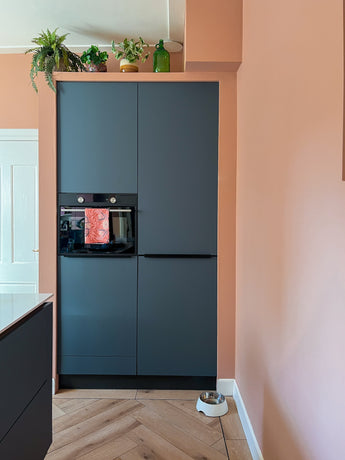 Un coin cuisine moderne avec des armoires sombres mates, un four encastré et un parquet clair. La plante artificielle Monstera 18 cm et une bouteille en verre verte se trouvent au-dessus des armoires ; un bol de nourriture pour animaux de compagnie se trouve sur le mur de pêche.