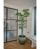 La plante artificielle Monstera 180 cm, une plante en pot haute avec de grandes feuilles fendues, est située dans une pièce moderne et lumineuse à côté d'une porte vitrée, d'un parquet, d'un canapé et de lattes verticales bleues.