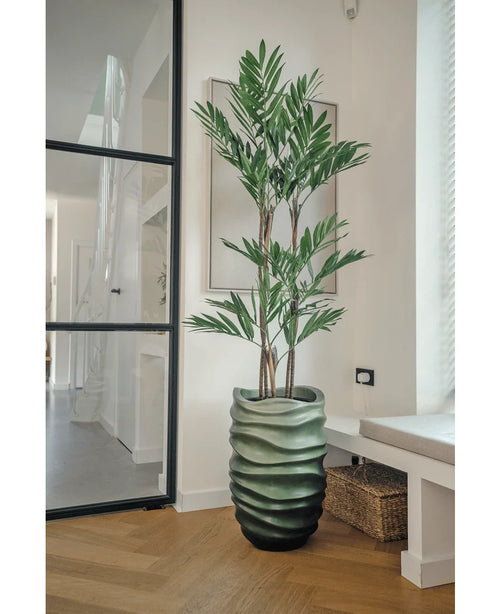 La plante artificielle Parlor Deluxe 120 cm a des tiges fines et de longues feuilles vertes dans un vase vert ondulé, parfait pour les pièces modernes et lumineuses avec des parquets en bois clair et de grandes fenêtres.