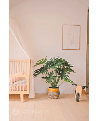 Une chambre d'enfant confortable et minimaliste avec un berceau et un auvent en bois à gauche, une plante artificielle Philodendron UV 90 cm au milieu comme pièce maîtresse, un vélo jouet en bois à droite et une simple impression d'art au trait au-dessus.