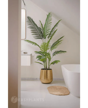 La plante artificielle Phoenix 180 cm dans un pot doré se trouve à côté d'une baignoire et d'un lavabo blancs dans une salle de bain moderne et minimaliste aux murs et au sol blancs. A proximité se trouve un tapis rond tissé sur le sol.