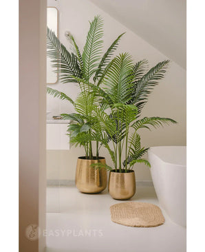 Une plante artificielle Phoenix de 180 cm avec des feuilles vertes luxuriantes dans un pot doré brillant se trouve à côté d'une baignoire blanche et d'un tapis tissé beige dans une salle de bain moderne et minimaliste, complétée par un miroir et une fenêtre pour un look serein.