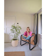 Une femme vêtue d'une chemise rose et d'un jean est assise pieds nus sur une chaise tressée, souriante et se relaxant dans une pièce lumineuse et minimaliste aux murs et au sol blancs. A côté d'elle se trouve la plante artificielle Ruscus de 120 cm.