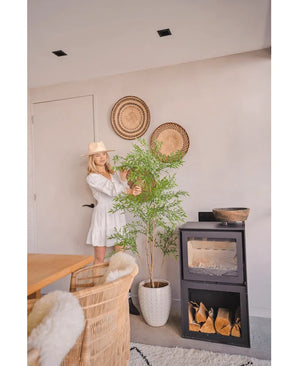 Une femme vêtue d'une robe blanche et d'un chapeau se tient à côté de la plante artificielle Ruscus de 150 cm, qui ajoute une touche naturelle à une pièce moderne avec des meubles en osier, des paniers muraux et un poêle à bois noir - parfait pour les intérieurs décoratifs.