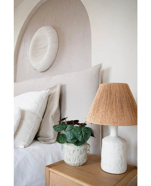 Une chambre moderne avec une palette de couleurs neutres comprend un lit avec une literie blanche, une table de chevet en bois, une lampe en céramique structurée avec abat-jour tissé, la plante artificielle Saxifraga de 27 cm et une sculpture murale ronde blanche au-dessus de la tête de lit.