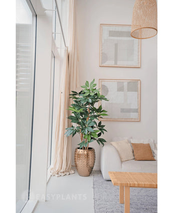 Een moderne, minimalistische woonkamer met grote ramen, een witte bank met beige kussens, een pot met gouden textuur met de Kunstplant Schefflera 150 cm, abstracte muurkunst en een geweven hanglamp boven je hoofd.